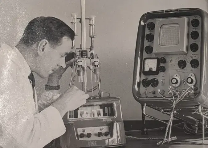 A black and white image of a male scientist using a microscope next to an old fashioned piece of electrical equipment