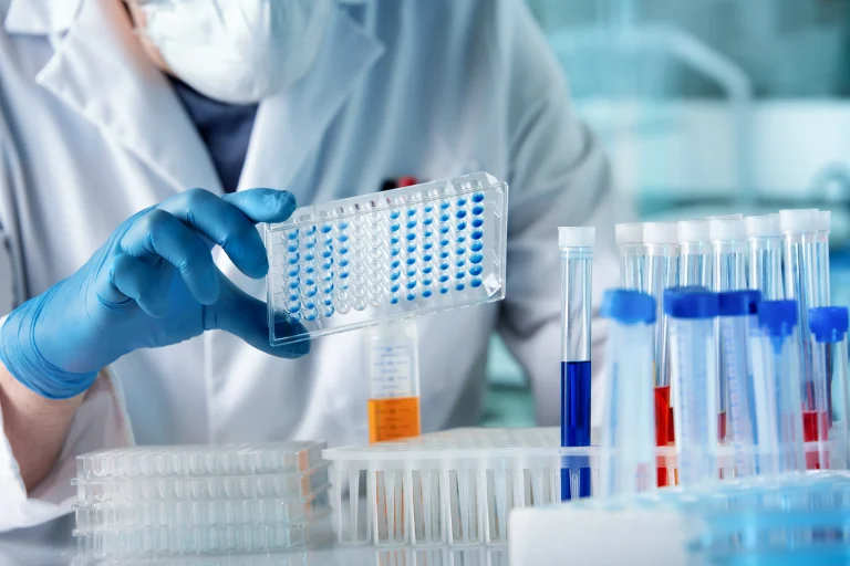 Lab technician in blue gloves holding a 96-well microplate with test tubes of colored samples in the foreground