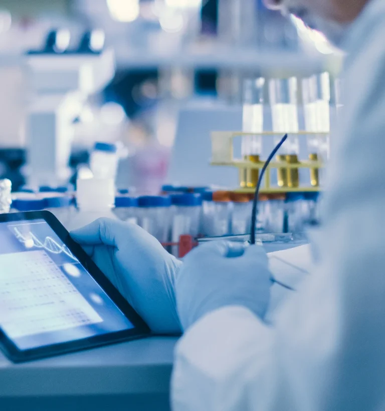 Gloved researcher reviewing data on a tablet in a laboratory, with test tubes and reagent bottles in the background