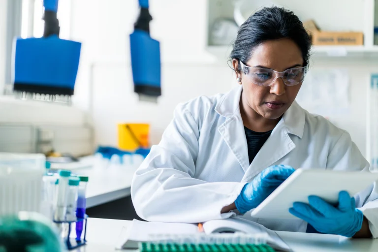 Researcher in safety glasses and gloves reviewing data on a tablet at a laboratory bench