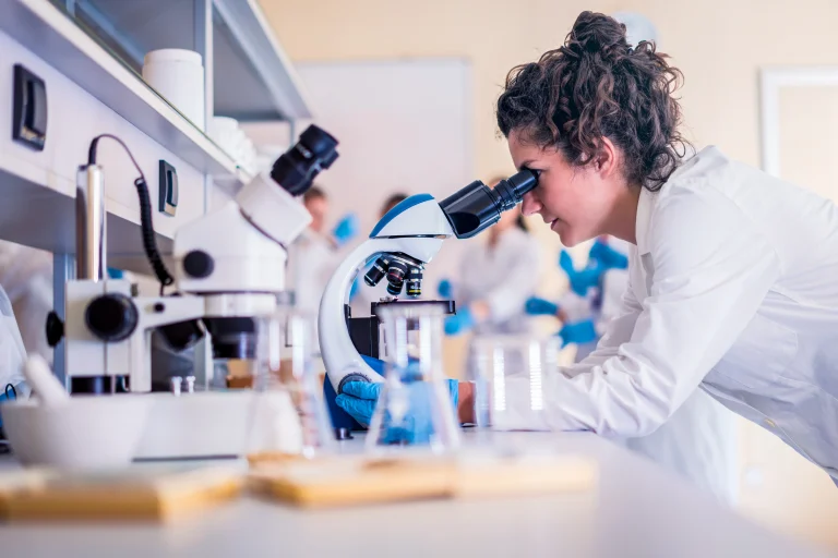 Scientist in a lab coat looking through a microscope in a clinical laboratory